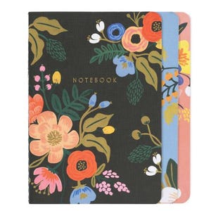 A set of three floral-patterned notebooks, each with a different colorful cover featuring vibrant flowers and leaves. The top notebook has \“NOTEBOOK\“ in gold letters on a dark background.