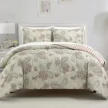 A white bed with a floral-patterned comforter set, including two matching pillow shams, is flanked by two white nightstands with lamps and decorative items. The wall features two framed prints and small windows, creating a cozy, coordinated bedroom look.