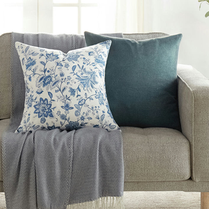 A light gray sofa features two decorative pillows: one with a blue floral pattern on white fabric and another in solid teal, complemented by a gray fringed throw draped over the arm.