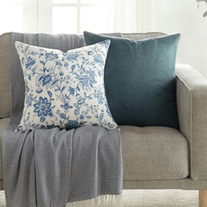 A light gray sofa features two decorative pillows: one with a blue floral pattern on white fabric and another in solid teal, complemented by a gray fringed throw draped over the arm.