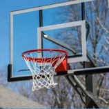 A Goliath brand basketball hoop with a clear glass backboard and red rim is mounted outdoors with a basketball net. The structure is set against a clear blue sky and bare trees.