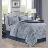 A bedroom scene showcasing a bed with a blue and white floral-patterned comforter set, two matching pillow shams, and a blue bed skirt. Flanked by two dark wood nightstands with lamps, it features a neutral headboard and framed floral artwork.