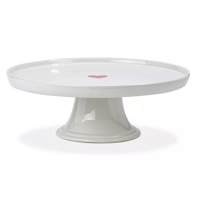 A white ceramic cake pedestal with a smooth finish, featuring a pink heart in the center. The pedestal has a round top and a sturdy base for supporting cakes or pastries.