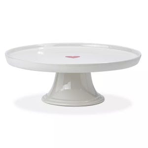 A white ceramic cake pedestal with a smooth finish, featuring a pink heart in the center. The pedestal has a round top and a sturdy base for supporting cakes or pastries.