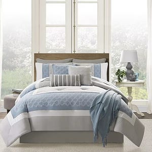 A bed with blue and grey bedding set including pillows, sheets, and a throw blanket in a room with a window.