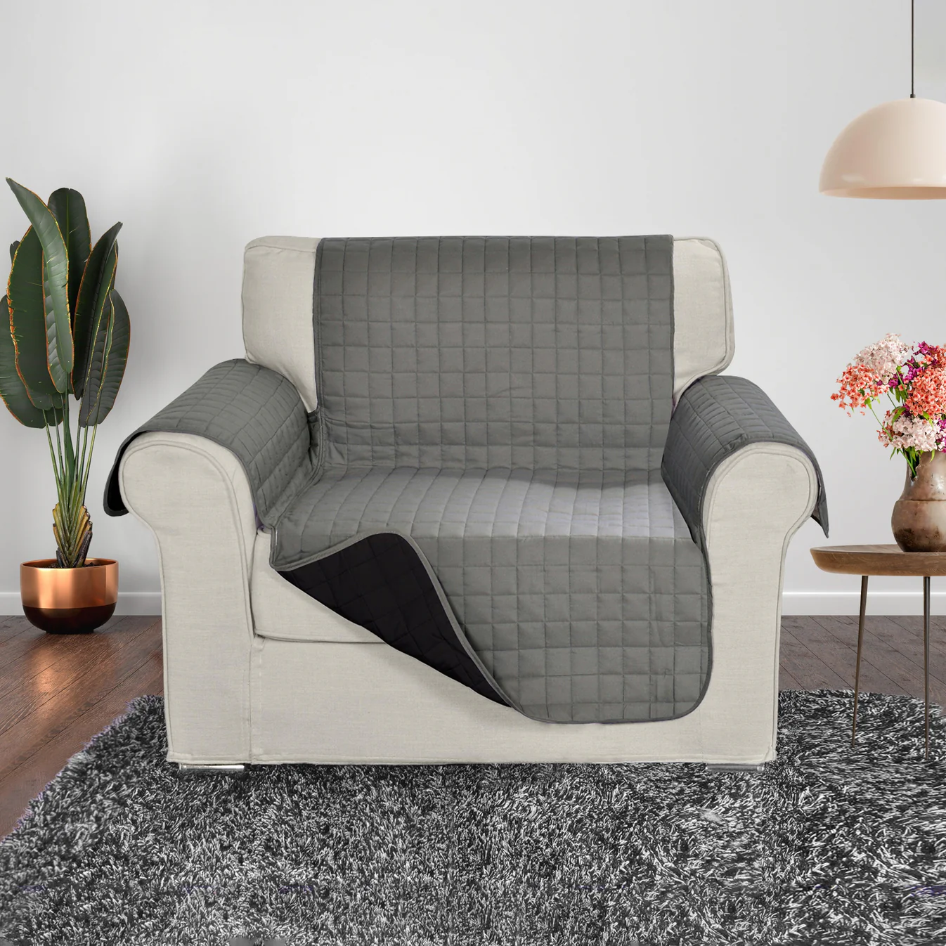 Armchair with a gray quilted cover, sitting on a gray shag rug. Nearby are a potted plant, a small table with flowers, and a hanging lamp.