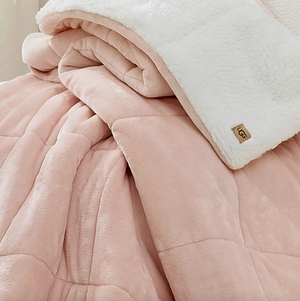 A soft pink quilted blanket with a fluffy white reverse side, featuring a small, tan \“UGG\“ label.