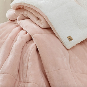 A soft pink quilted blanket with a fluffy white reverse side, featuring a small, tan \“UGG\“ label.