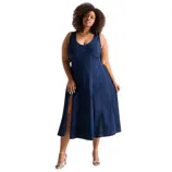 A woman is wearing a sleeveless, deep blue denim midi dress with a V-neckline and side slit, paired with silver strappy high-heeled sandals and a gold necklace.