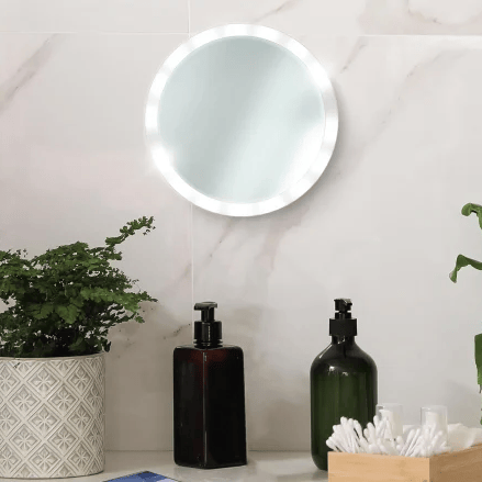 A round LED mirror is mounted on a wall with two soap dispensers, a plant in a patterned pot, and a wooden box with cotton swabs. A smartphone lays on the counter.