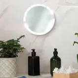 A round LED mirror is mounted on a wall with two soap dispensers, a plant in a patterned pot, and a wooden box with cotton swabs. A smartphone lays on the counter.