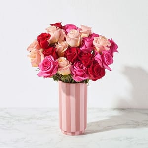 A bouquet of mixed roses in shades of red, pink, and peach is arranged in a pink striped vase set against a marble surface.