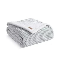 A textured throw blanket with a waffle-like design in a soft gray color, featuring a white Sherpa fleece underside and a small logo tag.