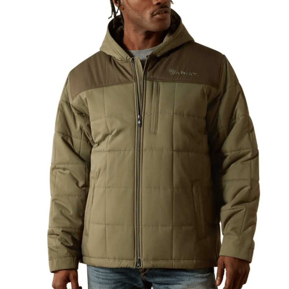 A man wearing an olive green Ariat padded jacket with a hood, featuring a front zipper and chest pocket, paired with blue jeans.