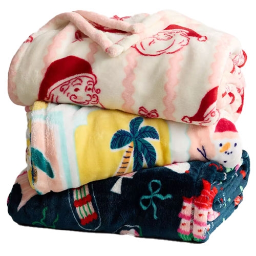 A stack of three folded blankets with festive designs: the top blanket features Santa faces, the middle one has palm trees and a snowman, and the bottom one displays holiday-themed patterns.