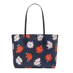 A navy tote bag with long handles featuring a floral pattern with red, white, and blue flowers. \“Kate Spade New York\“ is printed on the front.