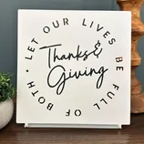 Decorative sign with the message: \“Let our lives be full of both Thanks & Giving,\“ featuring a stylized font. It includes a small plant and a wooden piece in the background.