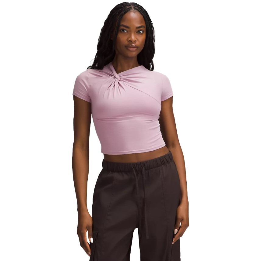 A woman is wearing a pink cropped top with a twist detail and high-waisted brown trousers.