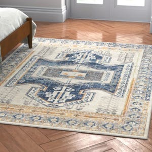 A rectangular area rug with a distressed geometric and floral pattern in blue, beige, and orange tones is placed on a herringbone wood floor beside a bed.