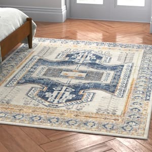 A rectangular area rug with a distressed geometric and floral pattern in blue, beige, and orange tones is placed on a herringbone wood floor beside a bed.