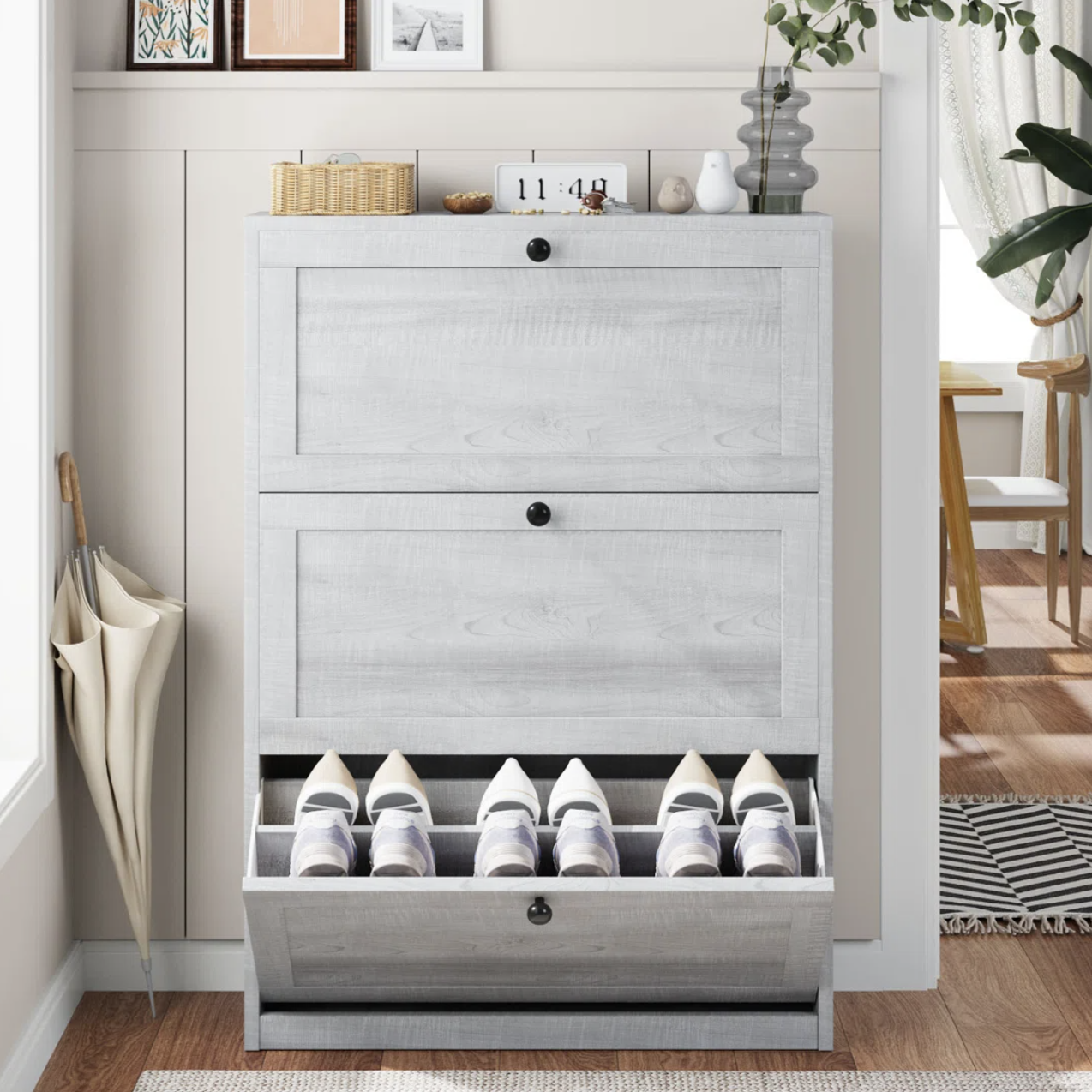 A light gray wooden shoe cabinet with three compartments holds multiple shoes, topped with decor items like framed pictures, a plant, and a clock. An umbrella rests beside it.