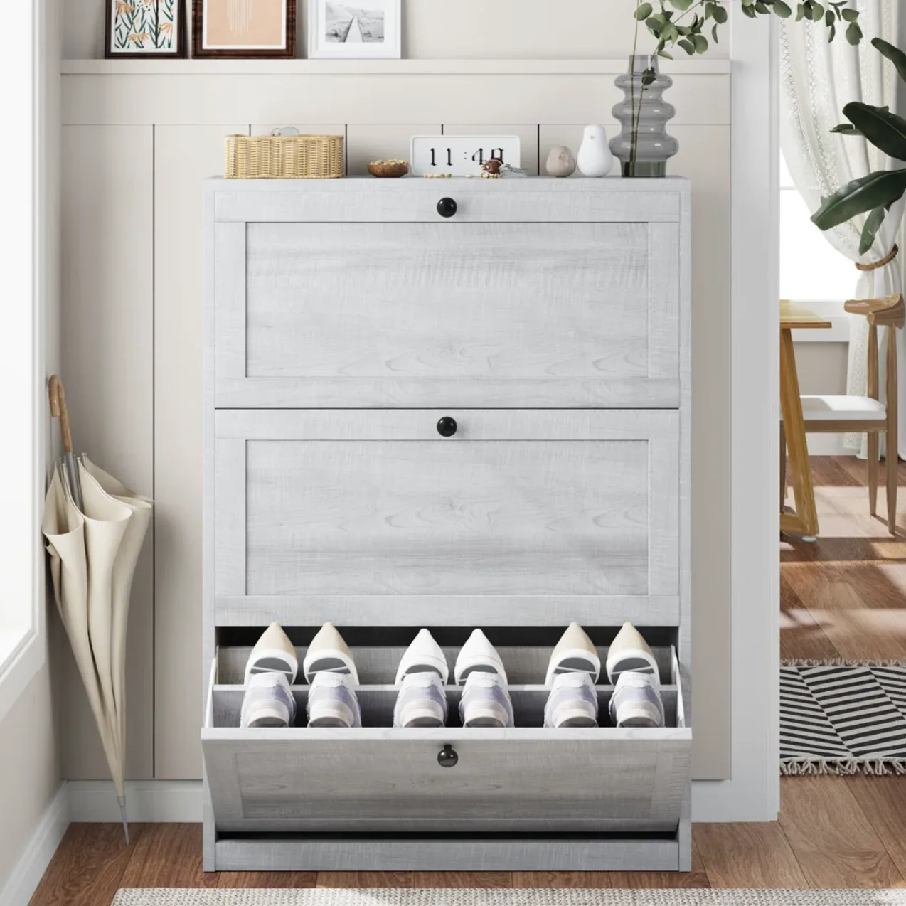 A light gray wooden shoe cabinet with three compartments holds multiple shoes, topped with decor items like framed pictures, a plant, and a clock. An umbrella rests beside it.