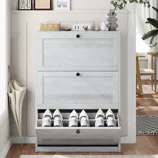 A light gray wooden shoe cabinet with three compartments holds multiple shoes, topped with decor items like framed pictures, a plant, and a clock. An umbrella rests beside it.