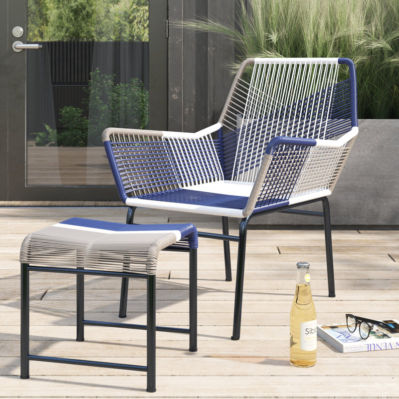A modern outdoor chair and ottoman set featuring a woven rattan design with blue, white, and gray tones on a black metal frame. They are placed on a wooden deck alongside a bottle of drink, glasses, and a magazine.