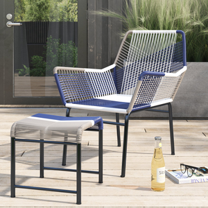 A modern outdoor chair and ottoman set featuring a woven rattan design with blue, white, and gray tones on a black metal frame. They are placed on a wooden deck alongside a bottle of drink, glasses, and a magazine.