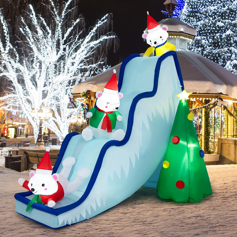 Inflatable Christmas decoration featuring three polar bears wearing Santa hats on a blue slide beside a Christmas tree with colorful ornaments, set in a snowy, illuminated winter scene.