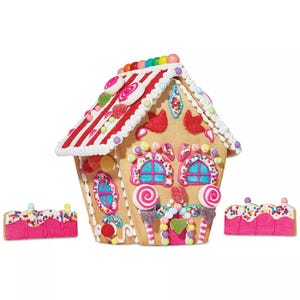Macy's DIY Holiday House Cookie Kit includes a gingerbread house decorated with colorful candy, icing details, and two pink rectangular cookies with sprinkles. The set is vibrant, featuring lollipops, jelly beans, and a variety of sweets for decoration.
