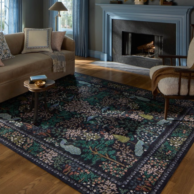 Living room with a beige sofa, blue-framed fireplace, blue curtains, patterned rug with nature motifs, wooden side table, and a cushioned armchair. Decor includes a lamp and wall sconces.