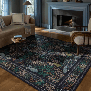 Living room with a beige sofa, blue-framed fireplace, blue curtains, patterned rug with nature motifs, wooden side table, and a cushioned armchair. Decor includes a lamp and wall sconces.