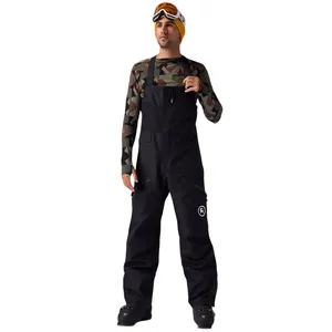 A person wearing camo-patterned long sleeves, black snow bib overalls, and ski boots, accessorized with an orange beanie and ski goggles.