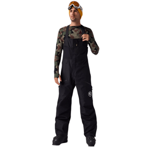 A person wearing camo-patterned long sleeves, black snow bib overalls, and ski boots, accessorized with an orange beanie and ski goggles.