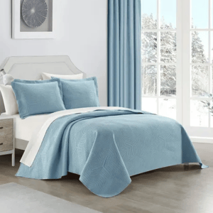 A bed is made with a light blue quilt and matching pillows, complemented by a white undersheet. A side table with a lamp is nearby, and the room features blue curtains and a snowy window view.
