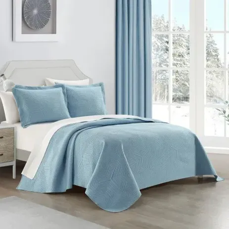 A bed is made with a light blue quilt and matching pillows, complemented by a white undersheet. A side table with a lamp is nearby, and the room features blue curtains and a snowy window view.