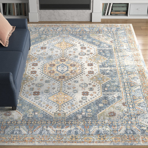 A machine-washable area rug measuring 8' x 10' features a geometric pattern with hexagonal shapes and floral motifs, primarily in blue, beige, and yellow tones, placed on a hardwood floor next to a navy couch.