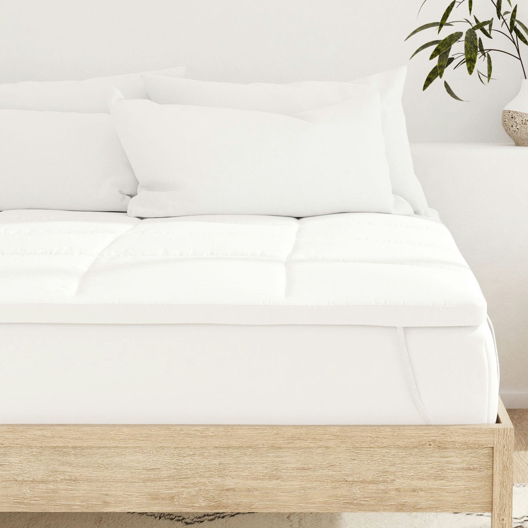 A neatly made bed with a white mattress topper, wooden frame, and several white pillows. A partial view of a plant in a decorative vase is visible on the side.