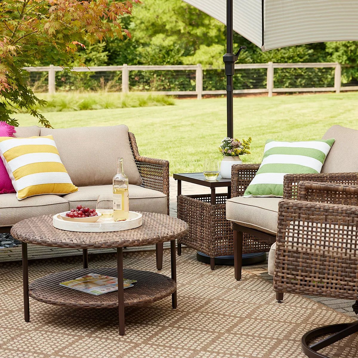 Outdoor patio set with wicker sofa and chairs on a checkered rug, featuring yellow and green striped pillows, a round wicker coffee table with a bottle, glass, and grapes on a tray, under a large umbrella.