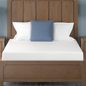 A wooden bed with a high headboard, white mattress, white pillows, and a blue decorative pillow is flanked by two wooden nightstands with modern lamps and a small plant.
