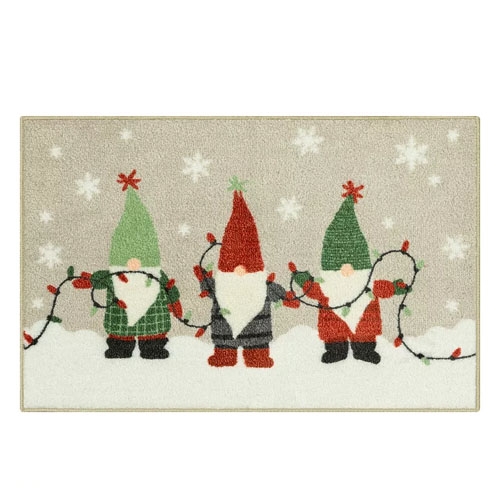 A decorative doormat featuring three gnomes holding colorful string lights, set against a snowy background with falling snowflakes.