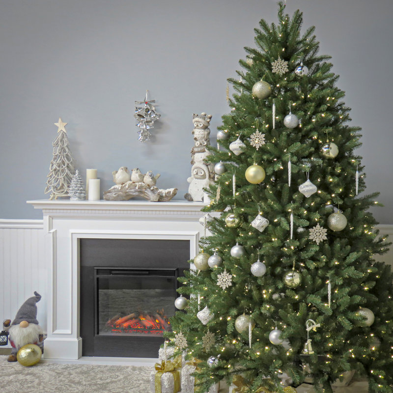 A decorated Christmas tree with silver and gold ornaments stands beside a white mantel with a faux fireplace. The mantel features bird figurines, candles, a decorative tree, and a small gnome.