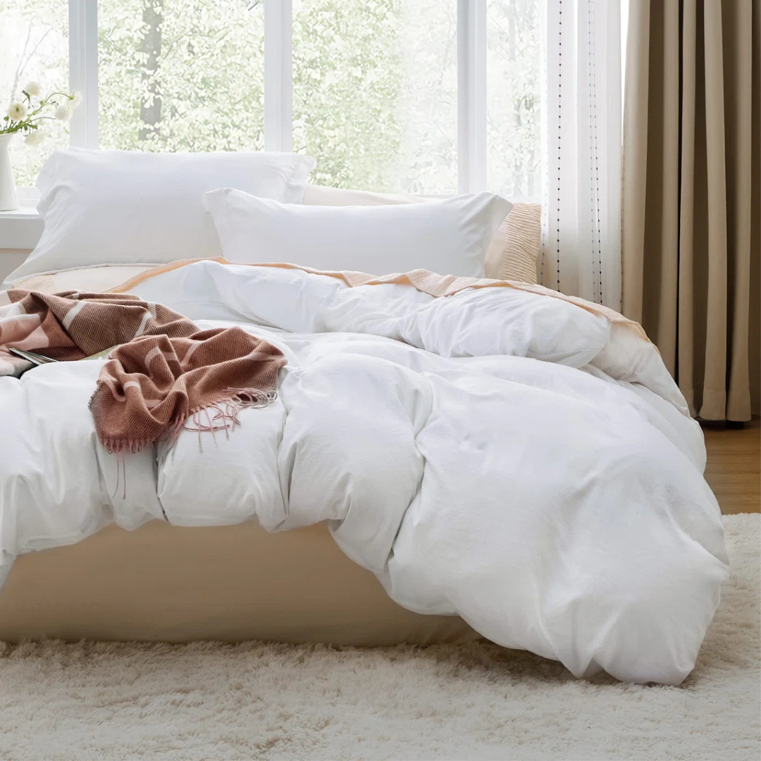 A cozy bed setup with fluffy white bedding and pillows, complemented by a soft, brown throw blanket. The room features a large window with curtains and a light-colored rug on the floor.