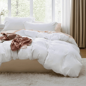 A cozy bed setup with fluffy white bedding and pillows, complemented by a soft, brown throw blanket. The room features a large window with curtains and a light-colored rug on the floor.