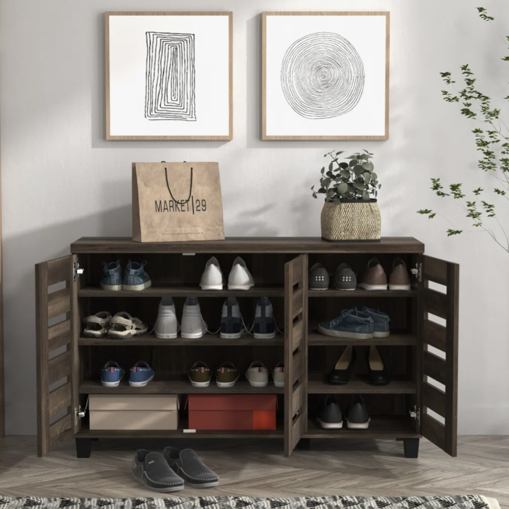 A wooden shoe cabinet with multiple shelves and two doors is filled with various shoes. A paper shopping bag, a potted plant, and two framed abstract artworks are placed on top, and a pair of slippers is on the herringbone-patterned floor, next to a patterned rug and nearby plant.