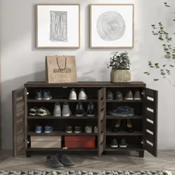 A wooden shoe cabinet with multiple shelves and two doors is filled with various shoes. A paper shopping bag, a potted plant, and two framed abstract artworks are placed on top, and a pair of slippers is on the herringbone-patterned floor, next to a patterned rug and nearby plant.