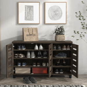 A wooden shoe cabinet with multiple shelves and two doors is filled with various shoes. A paper shopping bag, a potted plant, and two framed abstract artworks are placed on top, and a pair of slippers is on the herringbone-patterned floor, next to a patterned rug and nearby plant.