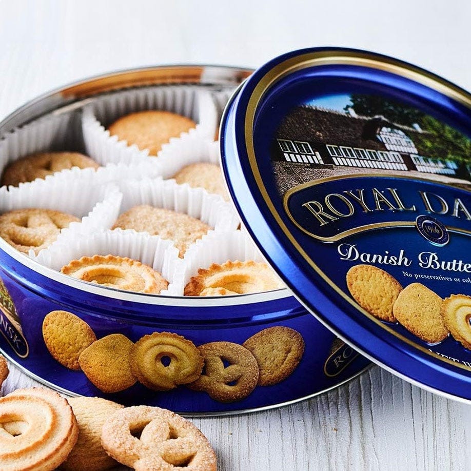 Blue tin of Royal Dansk Danish Butter Cookies, featuring an assortment of cookies in white paper cups with a variety of shapes like pretzels and swirls. The lid shows an image of a house and emphasizes no preservatives or colors are added.