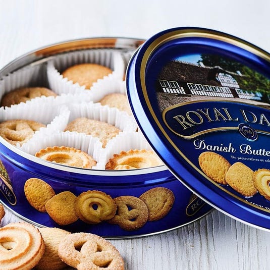 Blue tin of Royal Dansk Danish Butter Cookies, featuring an assortment of cookies in white paper cups with a variety of shapes like pretzels and swirls. The lid shows an image of a house and emphasizes no preservatives or colors are added.